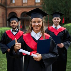 Graduate female shows like with her friends in graduation gowns holding diploma and smiling at camera.